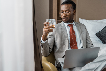 handsome african american businessman with laptop looking at glass of cognac in hotelの写真素材