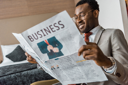smiling african american businessman in suit reading business newspaper in hotel roomの写真素材