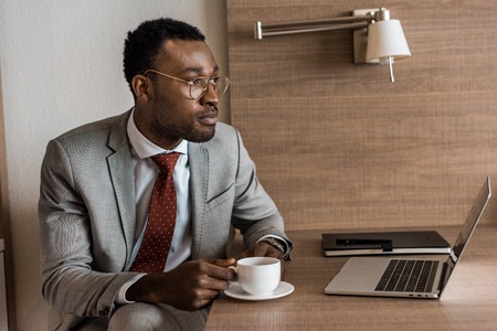 african american businessman holding cup of coffee at table with laptopの写真素材