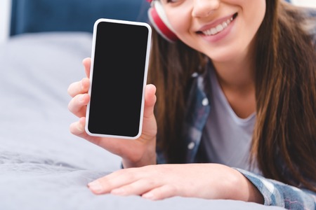 cropped shot of smiling young woman in headphones holding smartphone with blank screen while lying on bedの写真素材