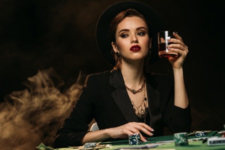 attractive girl in jacket and hat holding glass of whiskey at poker table in casino and looking awayの写真素材