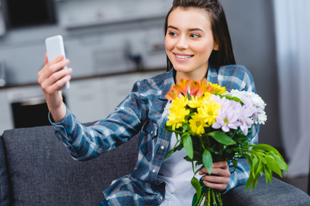 happy young woman holding beautiful flowers and taking selfie with smartphone at homeの写真素材