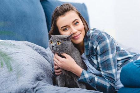 happy girl hugging cute grey cat and smiling at camera in bedroomの写真素材