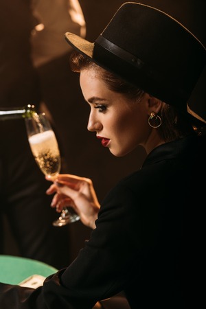 side view of waiter pouring champagne in glass of attractive girl in jacket and hat in casinoの写真素材