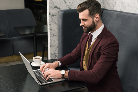 handsome businessman sitting at table with cup of coffee and typing on laptop in restaurantの写真素材