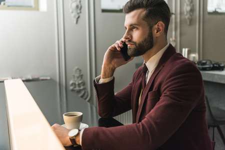 handsome businessman in formal wear sitting at table and talking on smartphone in restaurantの写真素材