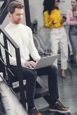 focused young man sitting in staircase and using laptop in officeの写真素材