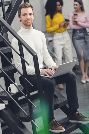 handsome man sitting with laptop on stairs and smiling at camera while female colleagues drinking coffee behind in officeの写真素材