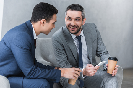 smiling businessmen drinking coffee from paper cups and using smartphone togetherの写真素材