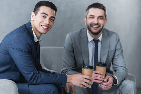 professional businessmen holding coffee to go and smiling at camera while sitting togetherの写真素材