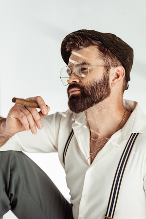 thoughtful bearded man in glasses and cap holding cigar on white backgroundの写真素材