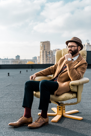 thoughtful businessman sitting in armchair on roof with buildings at backgroundの写真素材