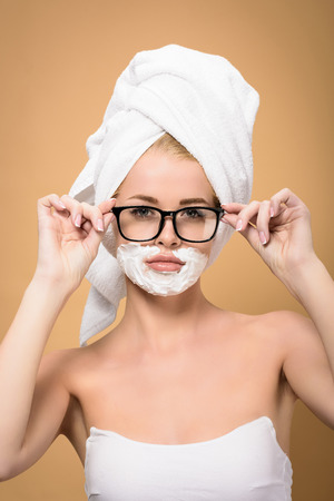 woman with towel on head and shaving cream on face adjusting eyeglasses and looking at camera isolated on beigeの写真素材