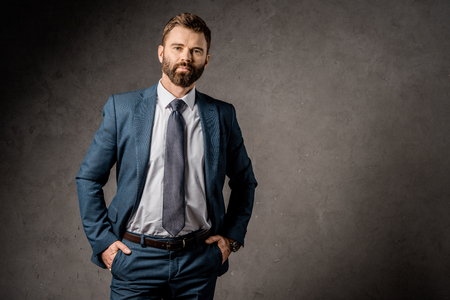 handsome bearded businessman standing in formalwear with hands in pocketsの写真素材