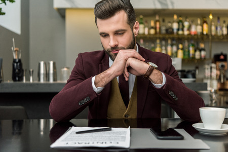 pensive businessman in formal wear with folded hands sitting at table and looking at contractの写真素材