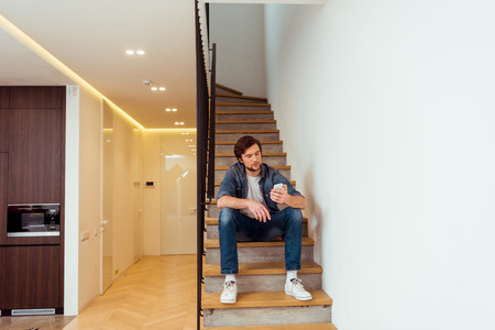 handsome man sitting on stairs and looking at smartphone screenの写真素材