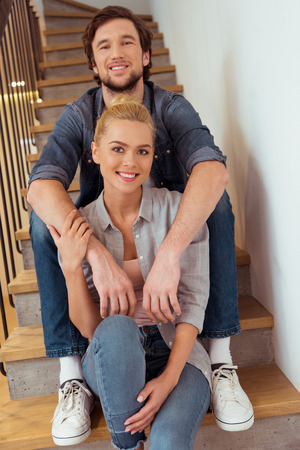 smiling man sitting on stairs and holding hands on shoulders of beautiful wifeの写真素材
