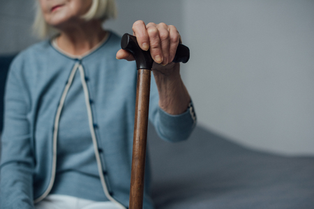 partial view of senior woman sitting on bed and holding walking stick at homeの写真素材