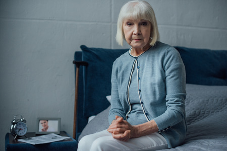 senior woman looking at camera and sitting on bed with folded hands at homeの写真素材