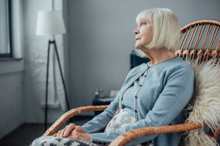 senior woman sitting in wicker rocking chair at home and looking awayの写真素材