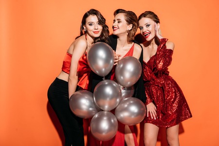 laughing attractive girls in stylish party clothes holding bundle of grey balloons isolated on orangeの写真素材