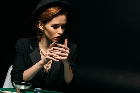 attractive girl in jacket and hat holding glass of whiskey at poker table in casino and looking awayの写真素材