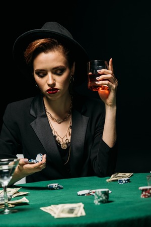 beautiful girl in jacket and hat holding glass of whiskey and poker chips at table in casinoの写真素材