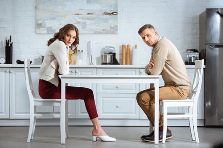 dissatisfied couple sitting with arms crossed at table in kitchenの写真素材