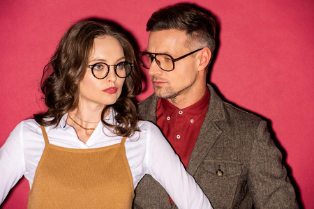 beautiful fashionable couple in formal wear and glasses posing on red backgroundの写真素材