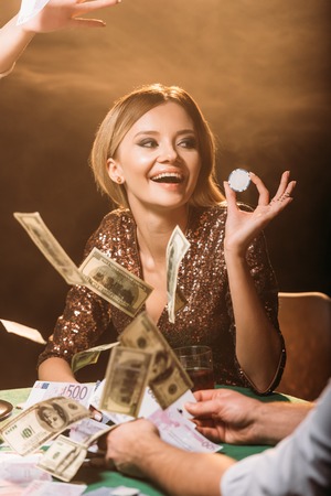 excited attractive girl holding poker chip with croupier at casino, dollar banknotes falling on...