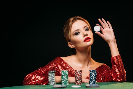 attractive girl in red shiny dress leaning on table, holding poker chip and looking at camera isolated on blackの写真素材