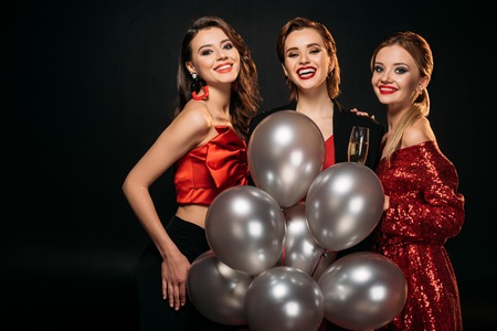 smiling attractive girls in stylish party clothes holding bundle of grey balloons and looking at camera isolated on blackの写真素材