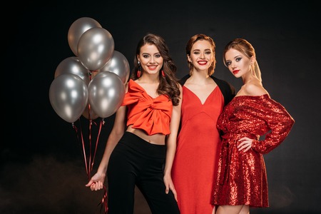 smiling attractive girls in red clothes holding bundle of grey balloons isolated on blackの写真素材