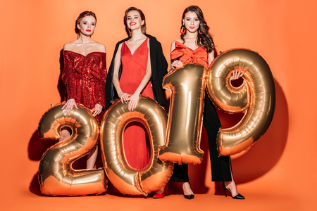smiling attractive girls in stylish party clothes holding 2019 balloons on orange, new year conceptの写真素材