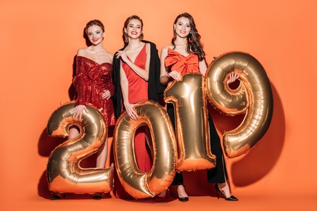 smiling attractive girls in stylish party clothes holding 2019 and looking at camera balloons on orangeの写真素材