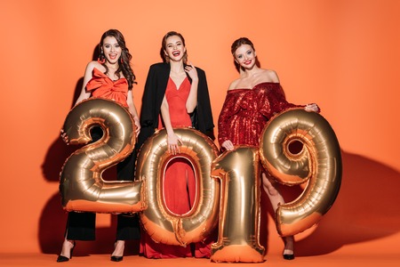 smiling attractive girls in stylish party clothes holding 2019 balloons on orangeの写真素材