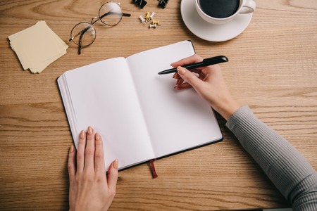 cropped view of businesswoman writing in notebook at workplace with glasses and cup of coffeeの写真素材