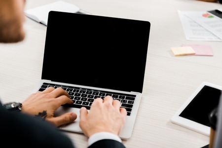 partial view of businessman typing on laptop with blank screen in officeの写真素材