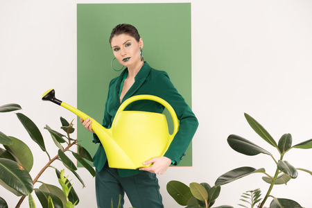 beautiful stylish woman holding watering can, looking at camera and posing with sea green and plants on backgroundの写真素材