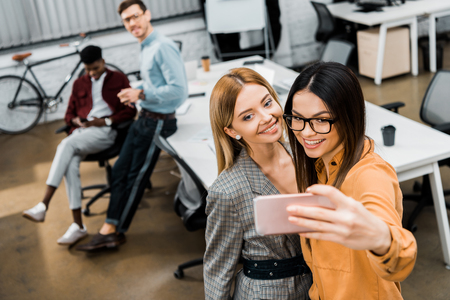 smiling businesswomen taking selfie on smartphone in office with multiracial colleagues behindの写真素材