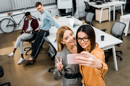 smiling businesswomen taking selfie on smartphone in office with multiracial colleagues behindの写真素材