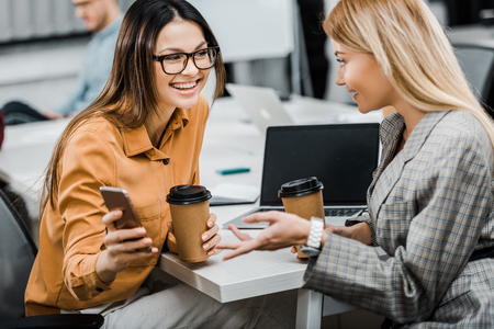 cheerful businesswomen using smartphone together at workplace in officeの写真素材
