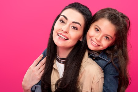 beautiful happy mother and daughter hugging and smiling at camera isolated on pink