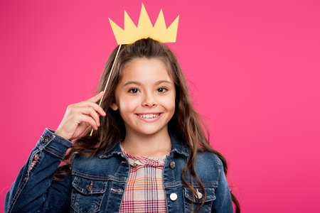 adorable happy child holding crown on party stick and looking at camera isolated on pinkの写真素材