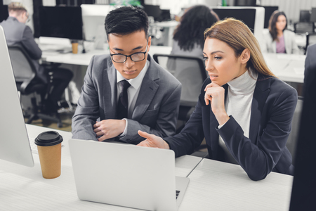 concentrated multiethnic businessman and businesswoman discussing work and pointing at laptop in officeの写真素材