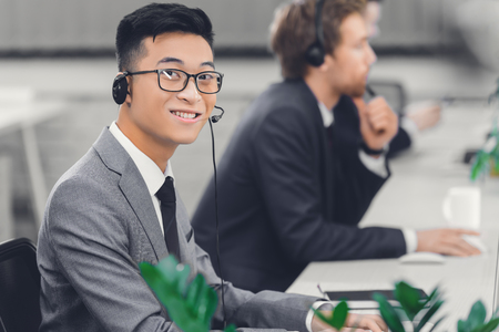 handsome young asian businessman in headset smiling at camera while working in call centerの写真素材