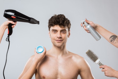 hands of hairstylists holding hairdryer, spray, comb and styling gel naer smiling man, isolated on greyの写真素材