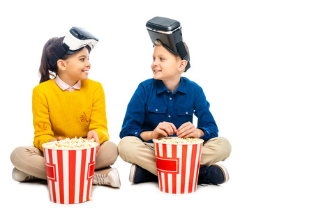 smiling kids with virtual reality headsets on heads sitting near striped popcorn buckets and looking at each other isolated on whiteの写真素材