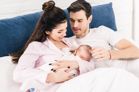portrait of young mother breastfeeding little baby with husband near by on bed at homeの写真素材