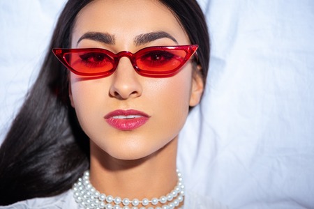 portrait of beautiful young woman in red eyeglasses lying on bed and looking at camera